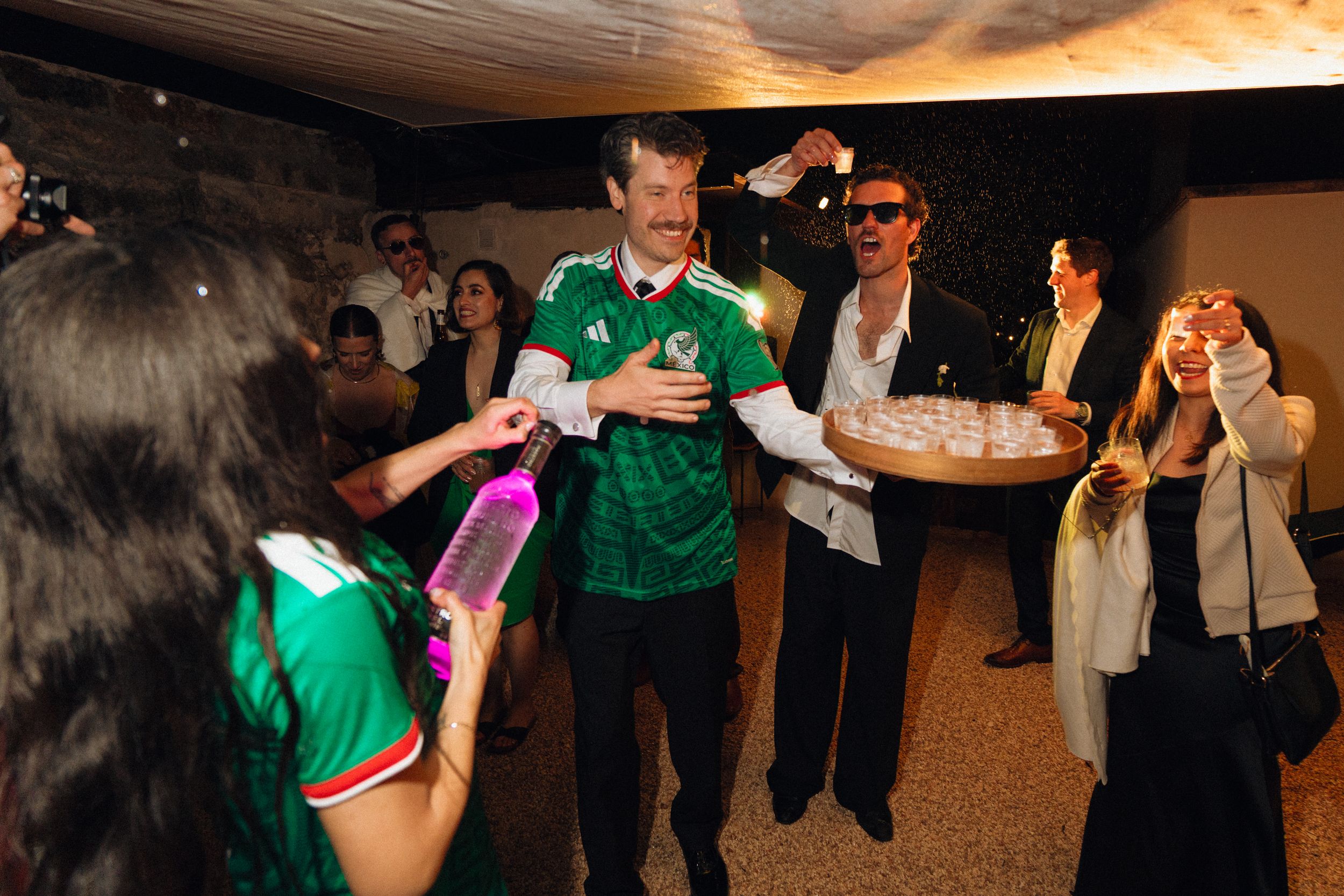People celebrating at a party, man in green Mexico soccer jersey surrounded by guests at an indoor event.