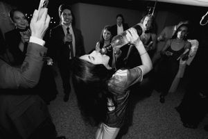 Black and white photo of a woman drinking from a bottle at a party with guests in formal attire.