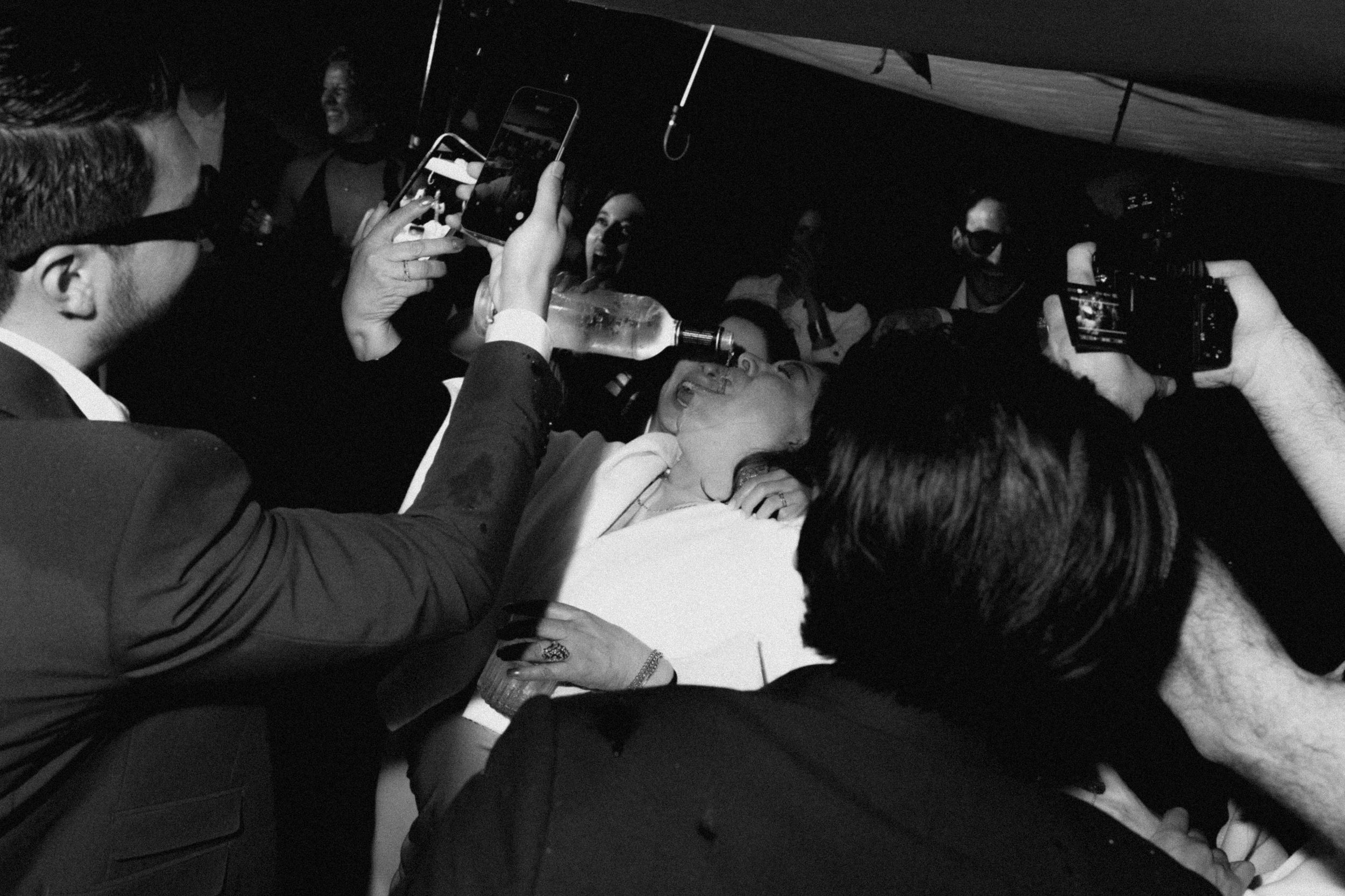 Black and white photo of a crowded event with people photographing a man in a white shirt surrounded by guests.