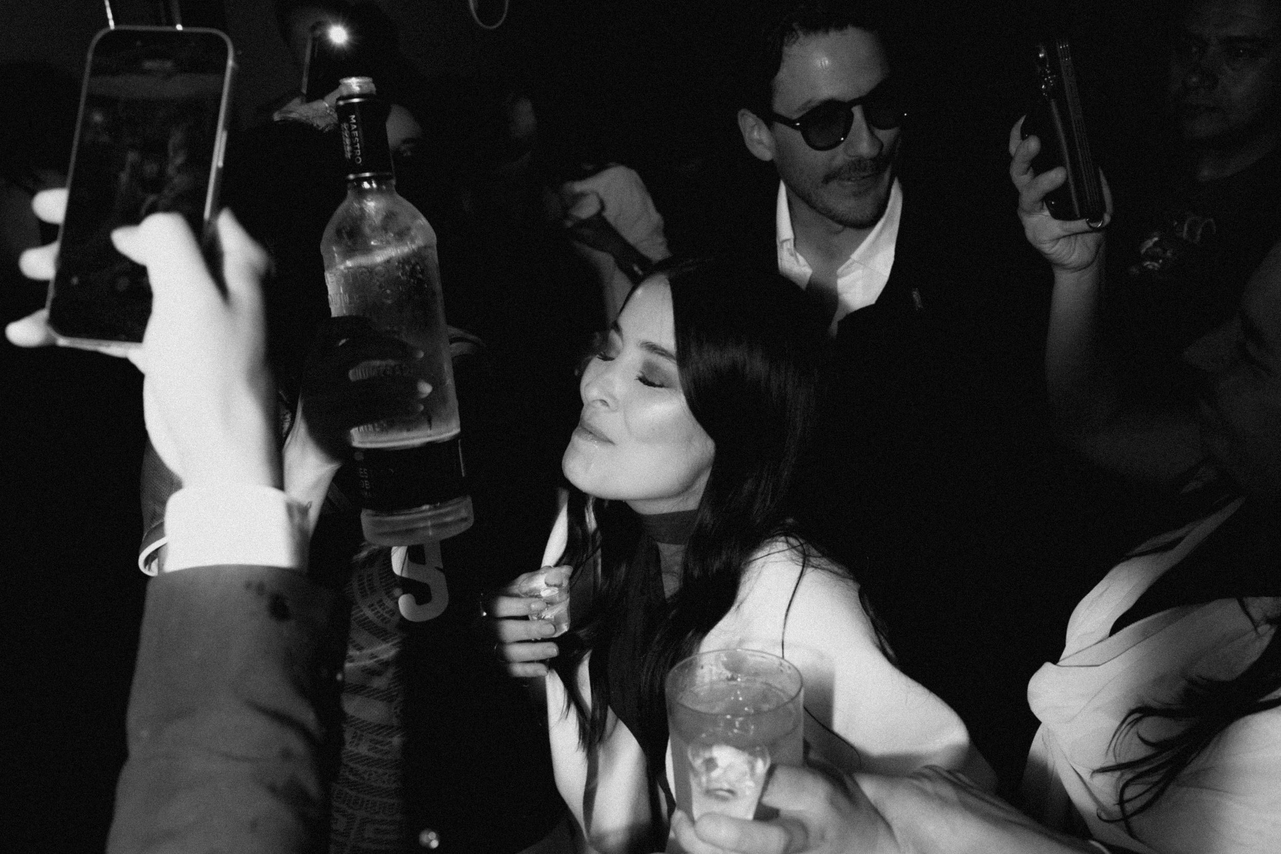 Black and white photo of a woman drinking from a bottle at a crowded party while guests take photos.