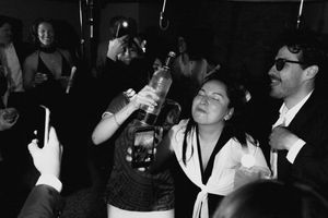 Black and white photo of people partying, woman drinking from bottle at lively social gathering.
