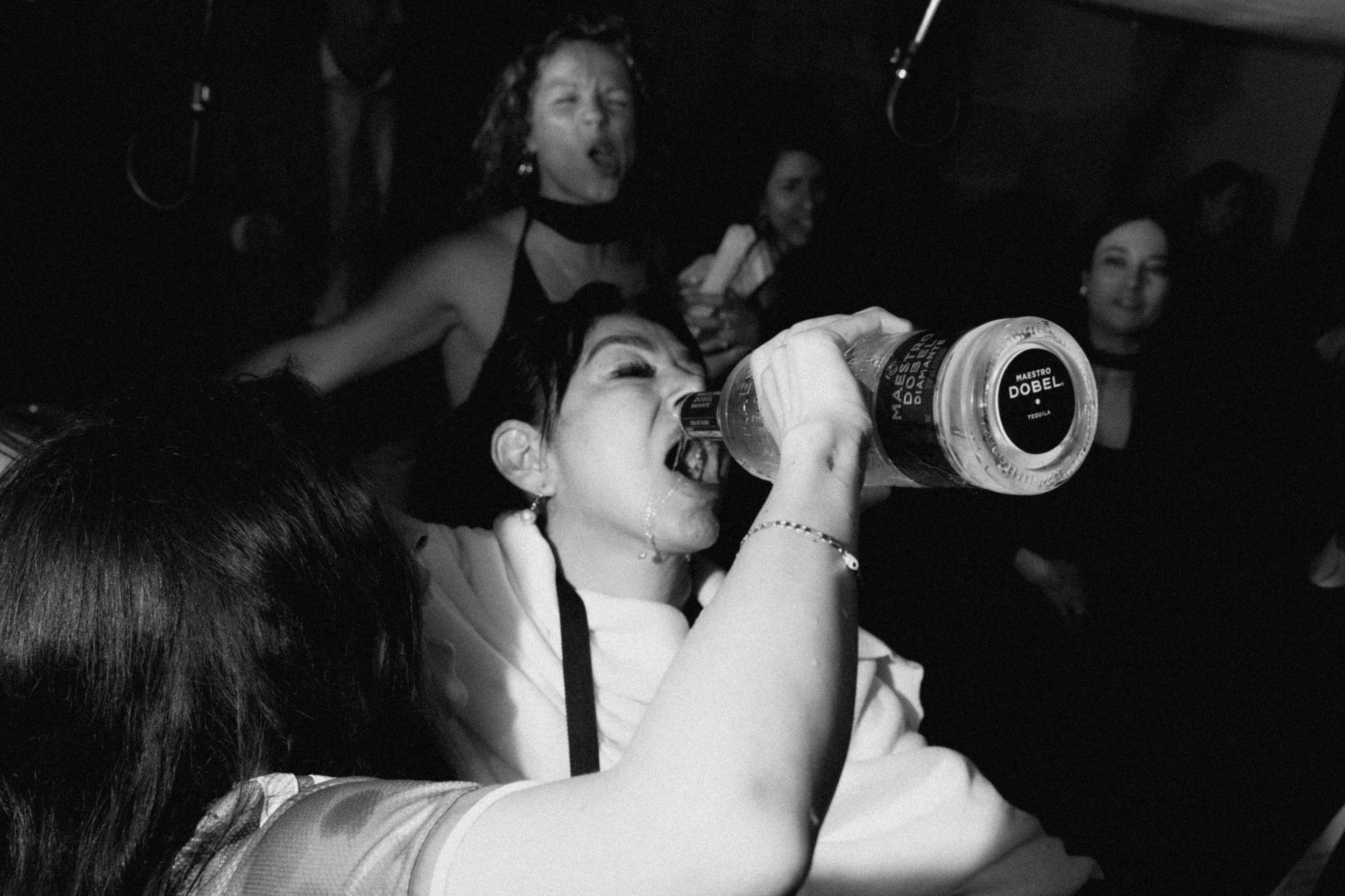 Black and white photo of a woman drinking from a bottle at a lively party.