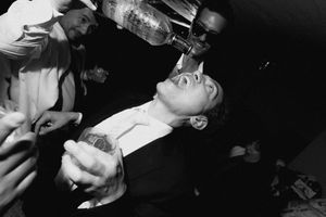 Black and white photo of a man having liquor poured into his mouth at a party.