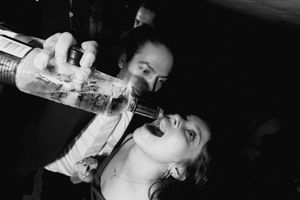 Black and white photo of a person pouring liquid from a bottle into another person's mouth at a party.