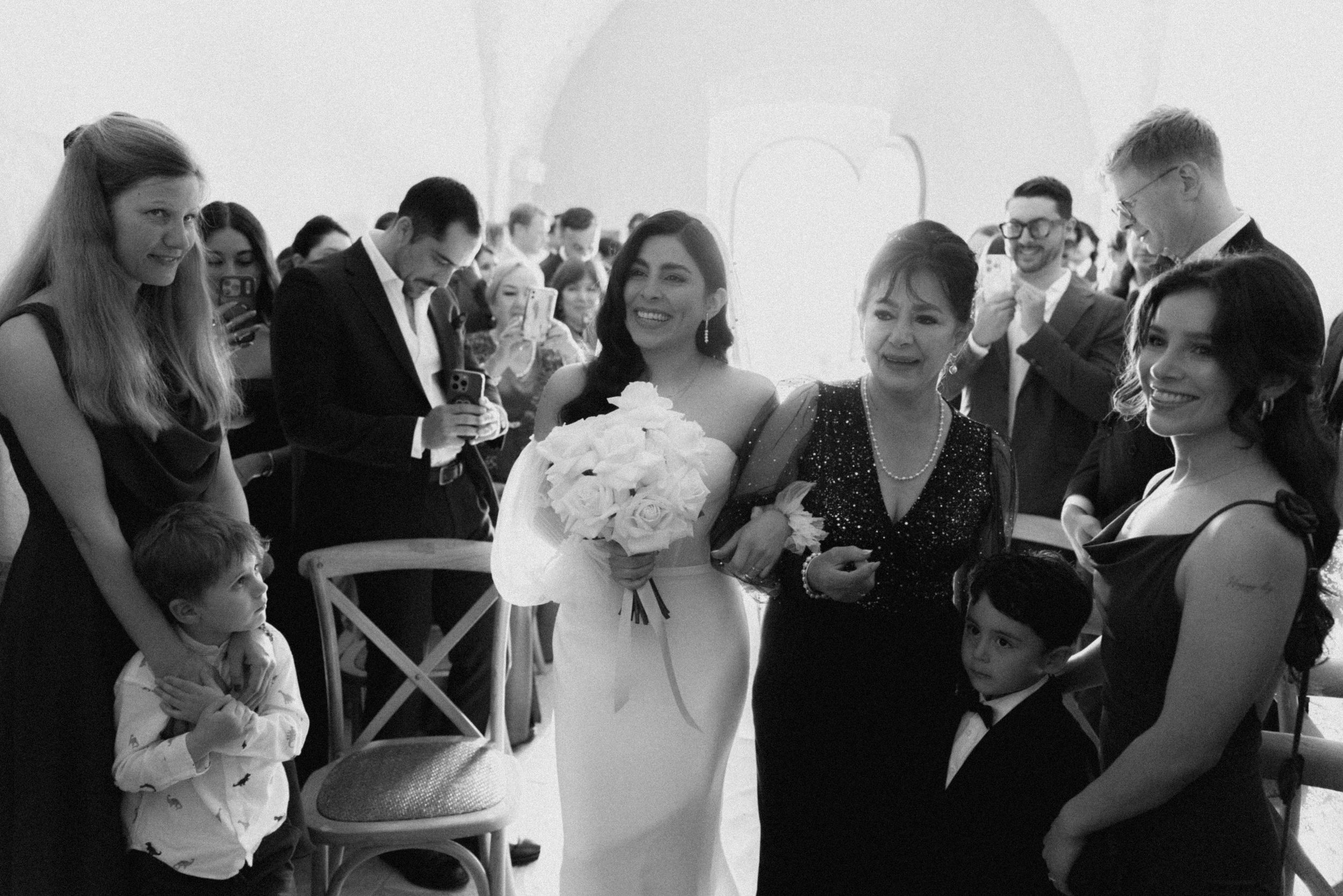 Bride walks down aisle holding white bouquet surrounded by smiling guests in black and white wedding photo.
