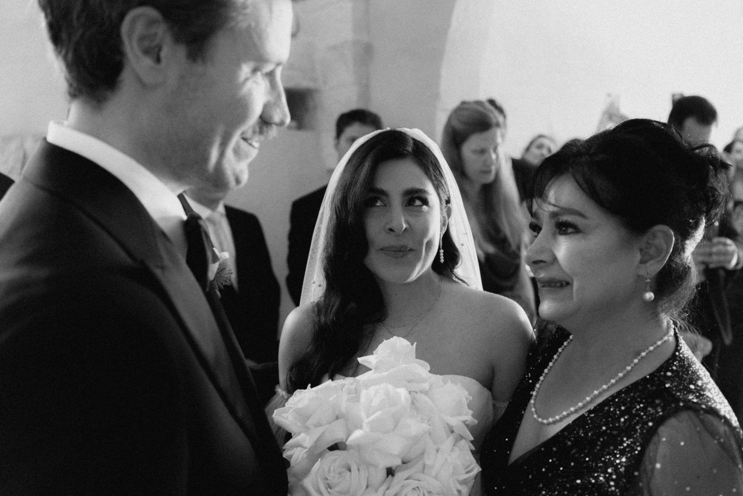 Black and white wedding photo of bride with bouquet, groom in tuxedo, and smiling woman in elegant gown.