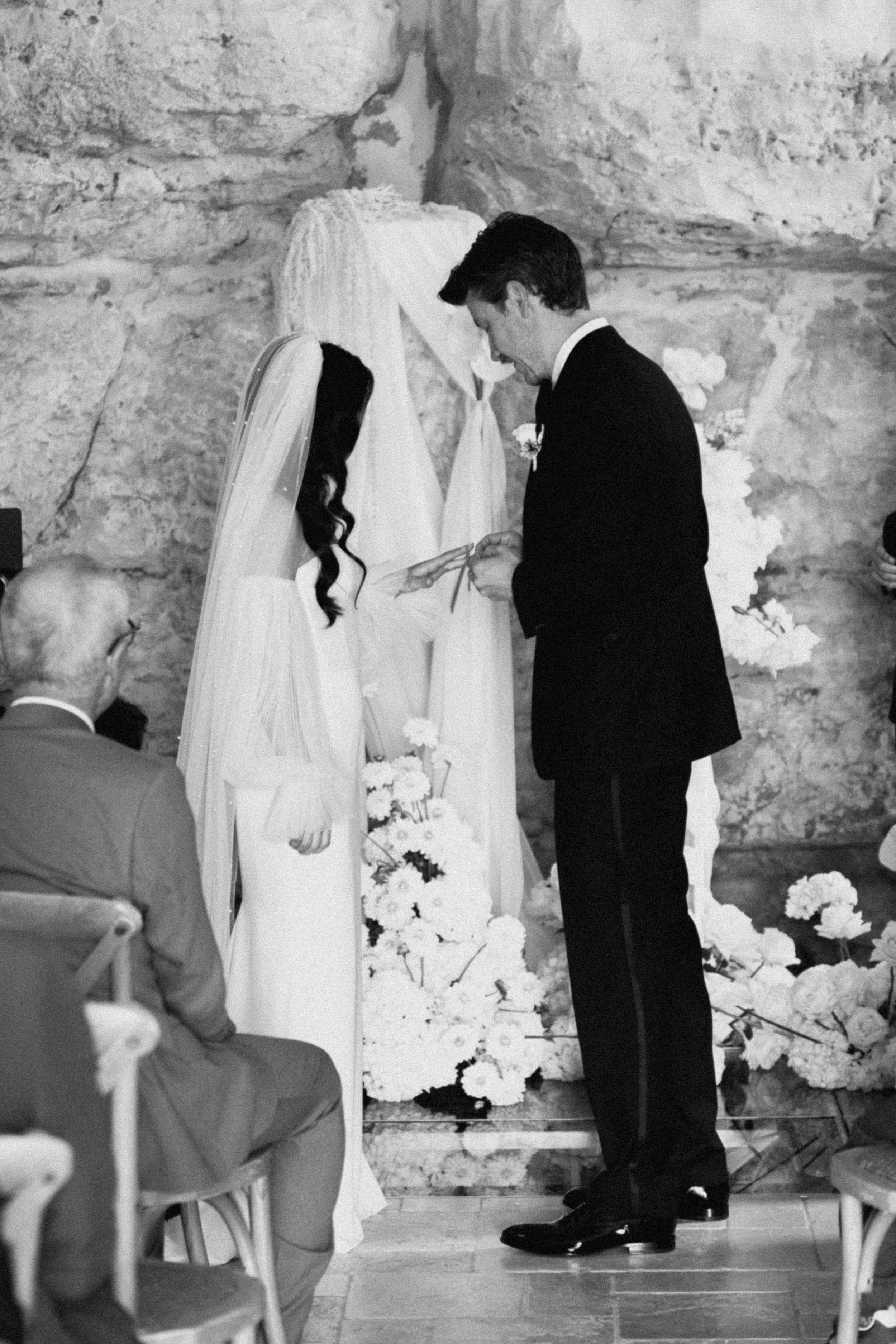 Black and white photo of bride and groom exchanging vows at elegant stone church wedding ceremony with floral arrangements.
