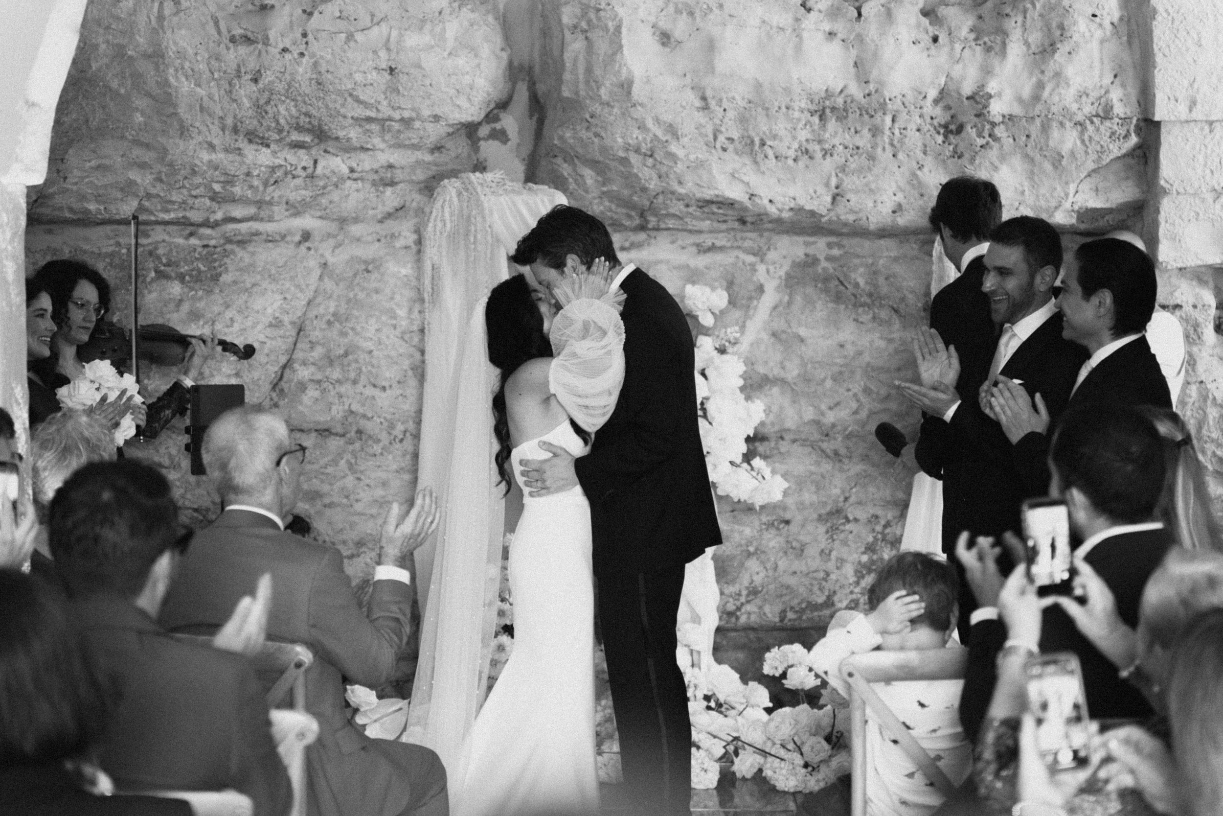 Black and white photo of a couple sharing their first kiss at a wedding ceremony surrounded by guests.
