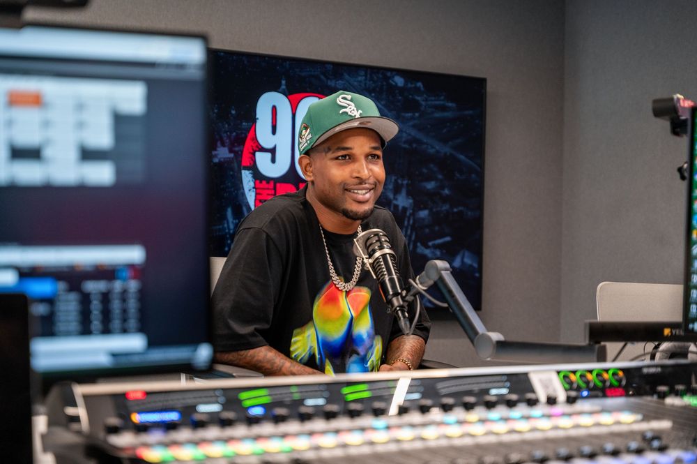 Man in black t-shirt and cap smiles at microphone in a professional radio studio with mixing board.