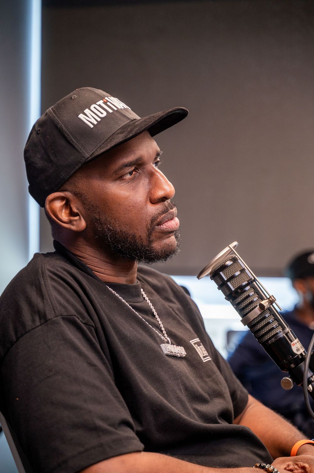 Man in black cap and t-shirt speaking into a studio microphone, wearing a chain necklace.
