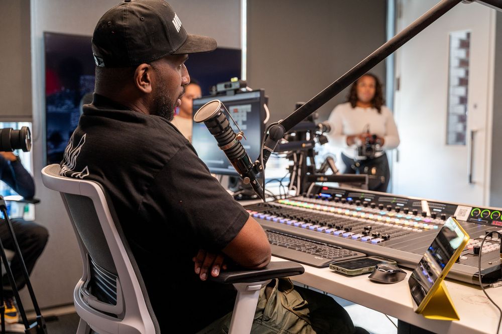 Man in black cap sits at a professional audio mixing console speaking into a microphone in a modern studio.