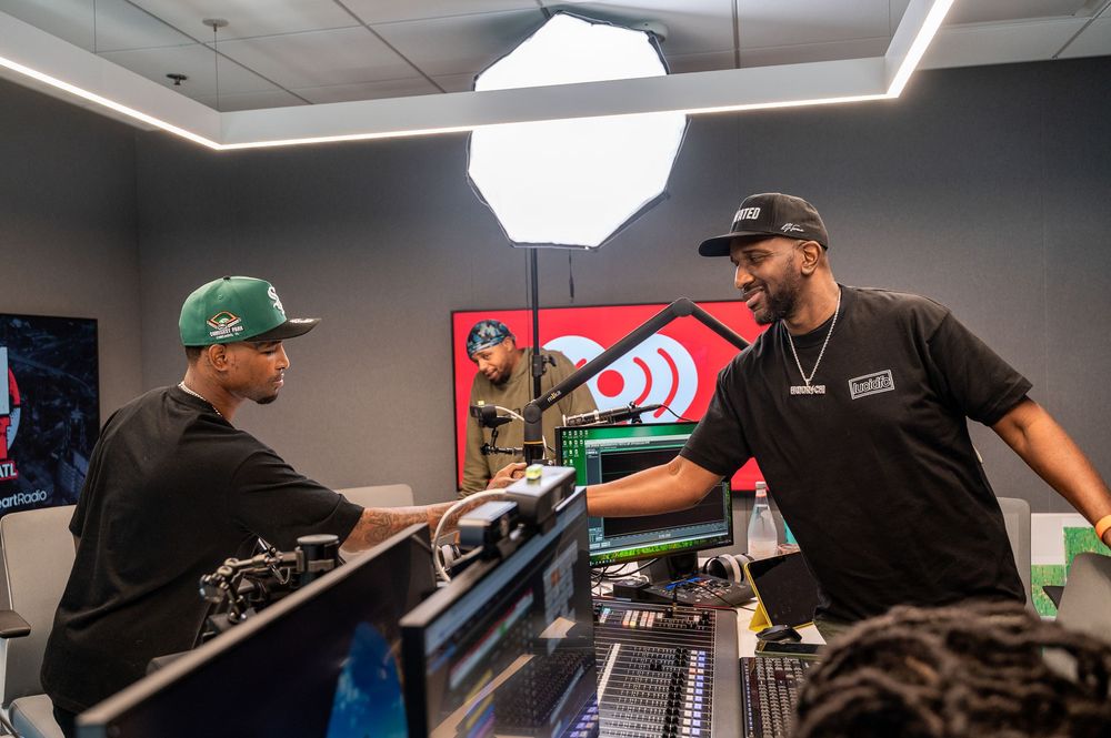 Two men collaborate in a professional music studio with mixing boards, bright lighting, and radio station branding visible.