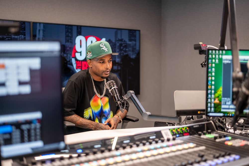 A rapper speaks into a microphone at a radio station studio with a mixing board in the foreground.