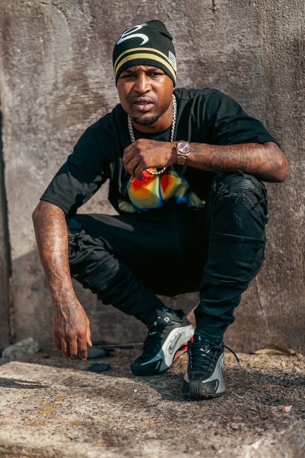 Man in black outfit and beanie crouching against concrete wall wearing chain necklace and sneakers.