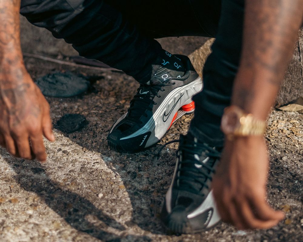 Close-up of a person wearing black Nike sneakers with red accents, crouching on rocky ground with tattooed arms.