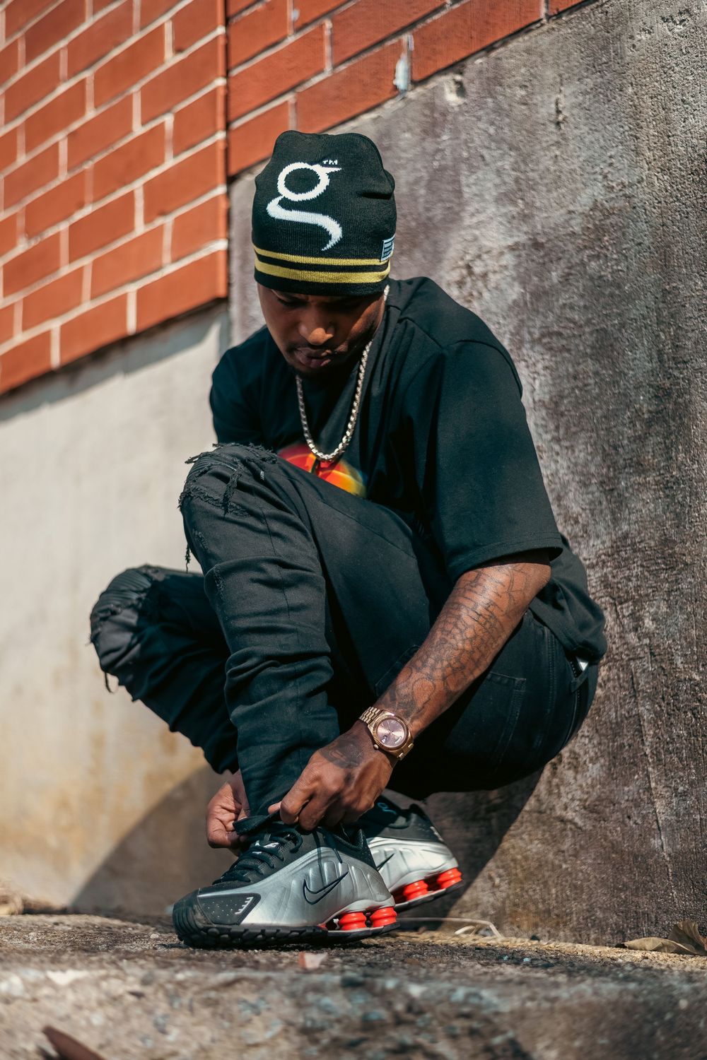 Man in black outfit and beanie tying silver and red Nike sneakers outdoors near brick wall.