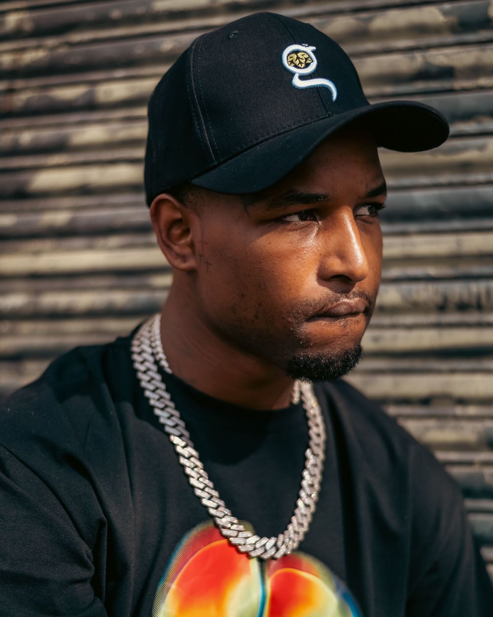 Man wearing black snapback cap and silver Cuban link chain posing outdoors against metal shutter backdrop.