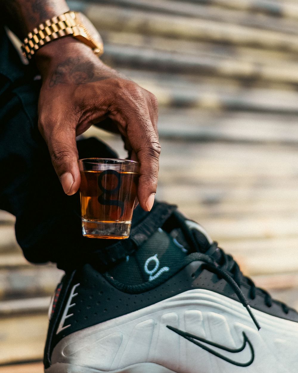 A hand holding a shot glass of whiskey next to a black and white Nike sneaker on wooden steps.