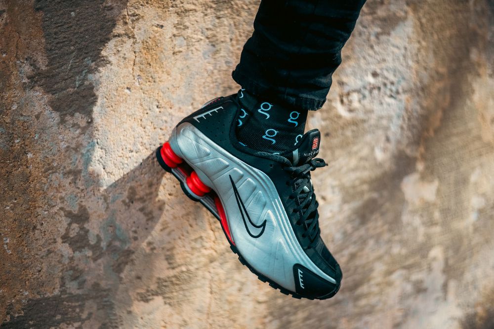 Nike Shox sneakers in black and red colorway worn with black pants against a textured concrete wall.