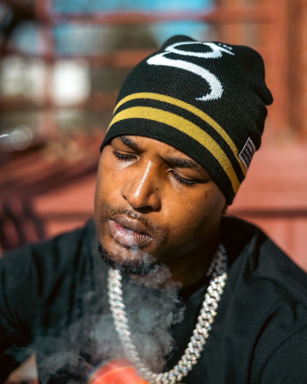 Man in black and gold beanie exhaling smoke wearing a silver chain necklace outdoors in sunlight.