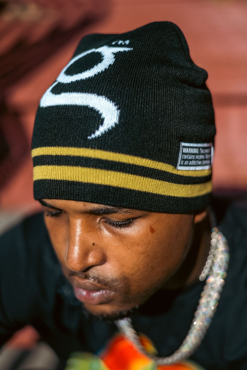 Young man wearing a black and gold beanie hat with white logo design, looking downward outdoors.