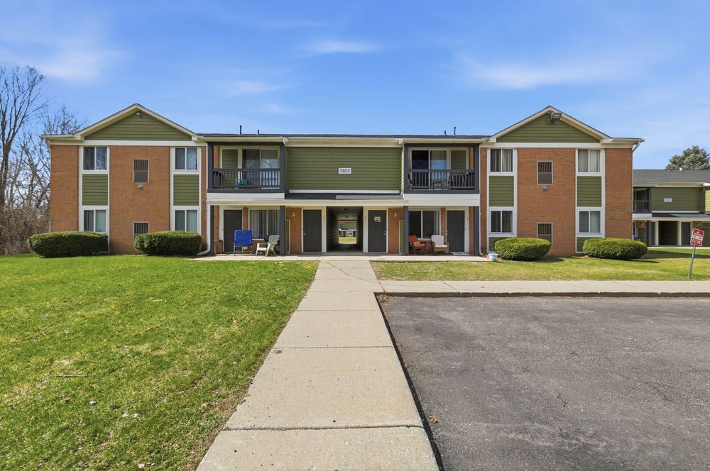 Two-story brick apartment complex with green trim, balconies, and manicured lawn on a sunny day.
