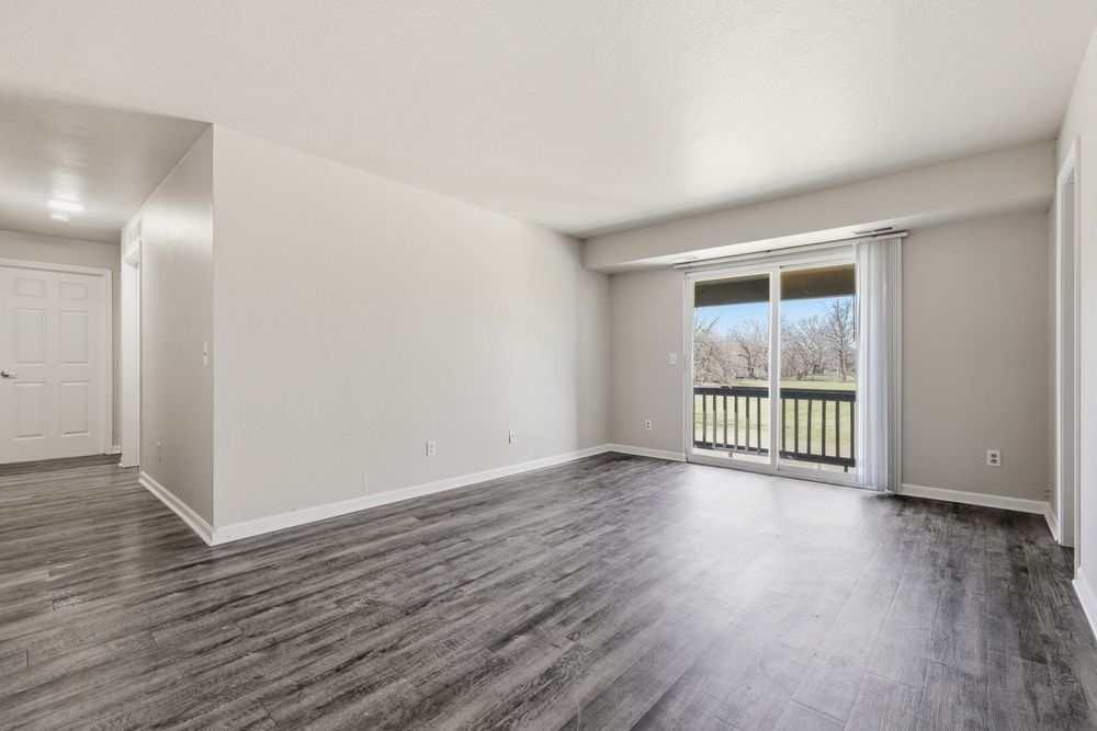 Empty living room with dark hardwood floors, white walls, and sliding glass door leading to balcony.