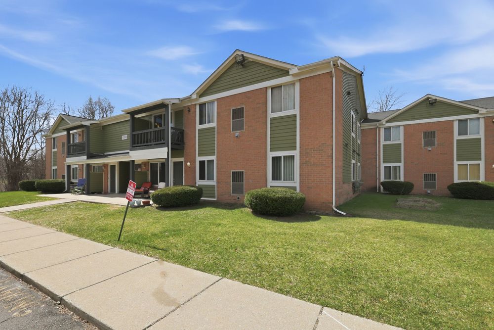 Brick apartment complex with green trim, manicured lawn, and sidewalk on a sunny day.