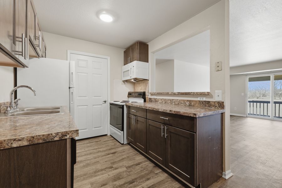 Modern kitchen with dark wood cabinets, granite countertops, white appliances, and hardwood floors leading to a bright balcony.