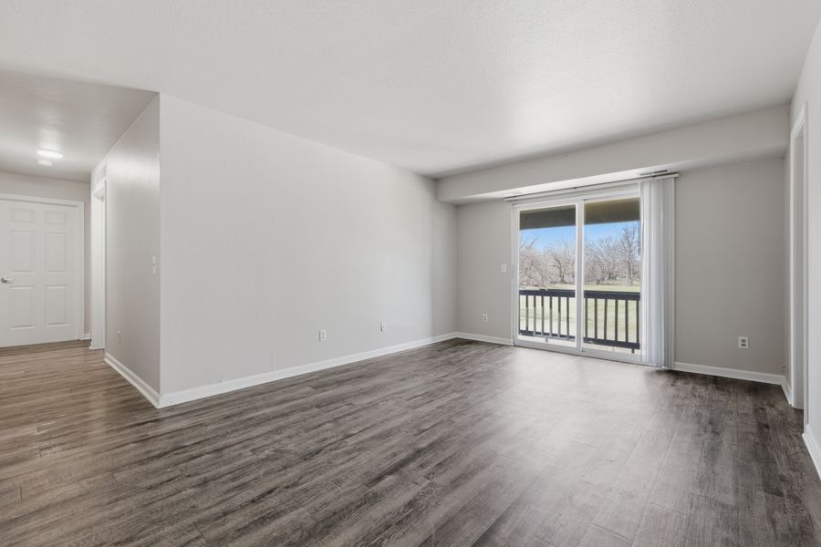 Empty living room with dark hardwood floors, white walls, and a sliding glass door leading to a balcony.