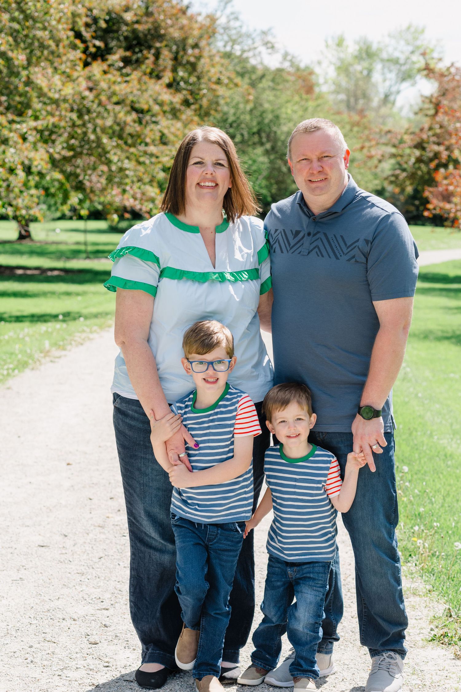 Happy family of four posing outdoors on a sunny day, with two young boys in striped shirts smiling in a park.