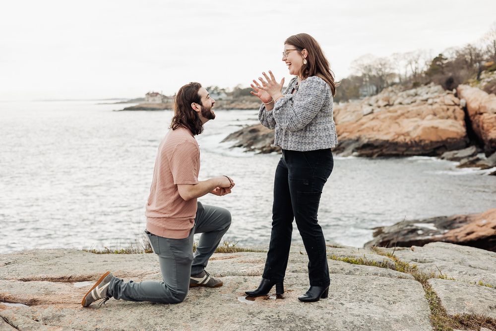 Man kneeling on rocky shore proposing to woman with ocean backdrop in a romantic outdoor marriage proposal scene.