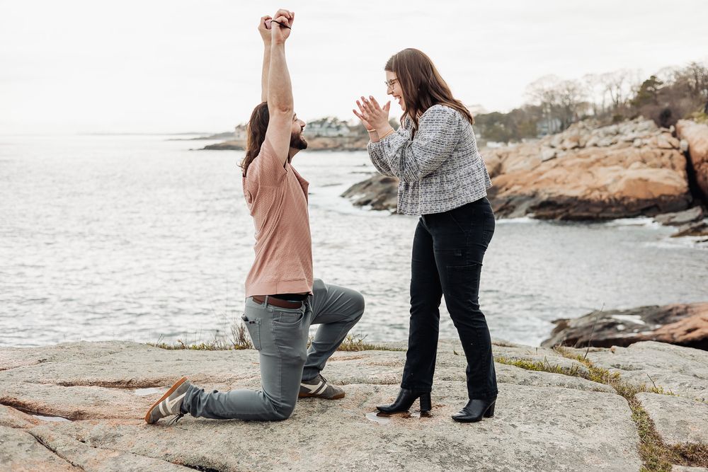 Same-sex couple proposal on rocky shoreline, one woman kneeling with ring raised while partner claps with joy.