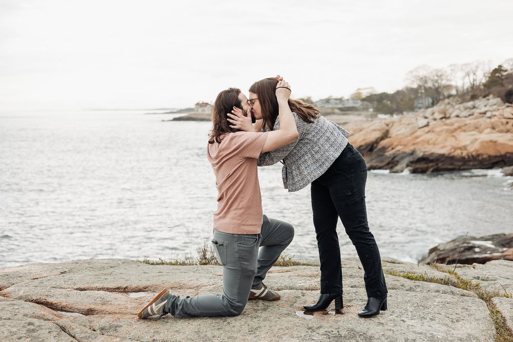 Couple sharing a romantic kiss during a seaside proposal on rocky New England coastline.