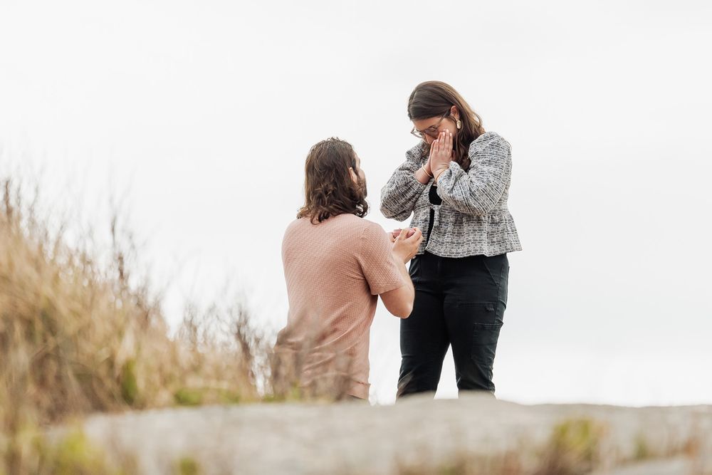 Person kneeling proposing to partner outdoors on rocky terrain with dry grass background on overcast day.