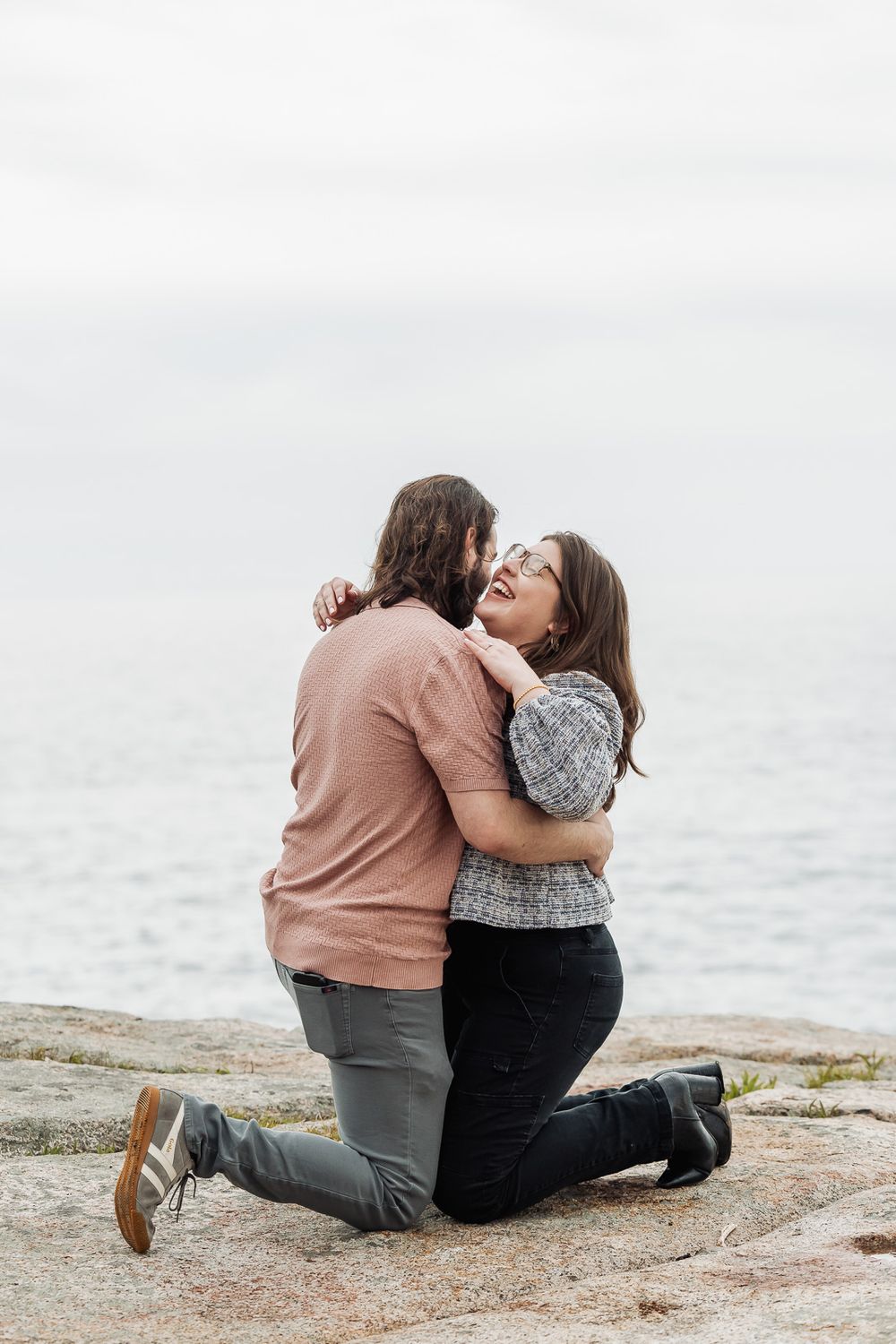 Couple embracing and laughing on rocky shore by calm water, woman in patterned top hugging man in pink shirt on knees.