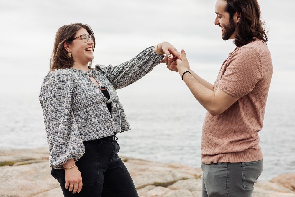 Couple holding hands and laughing together on rocky seaside, woman in grey jacket and man in pink shirt.