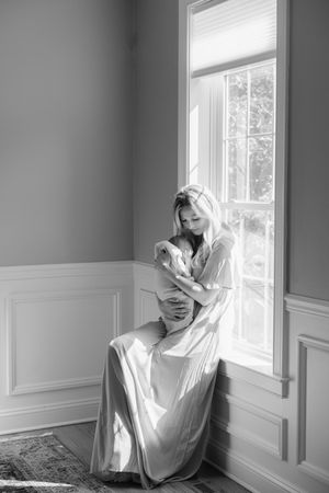 Black and white photo of a woman in a flowing gown cradling a newborn by a sunlit window.