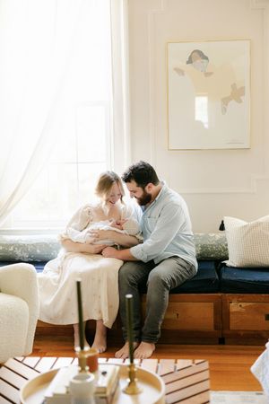 A couple sits by a bright window, tenderly gazing at their newborn baby in a cozy, elegantly decorated living room.