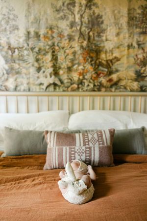 Elegant bed with rust linen, patterned pillows, a ceramic figurine, and a vintage floral tapestry on the wall.