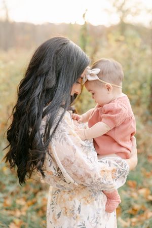 A mother with long dark hair holds her baby girl in a pink outfit during a golden hour outdoor autumn photo session.