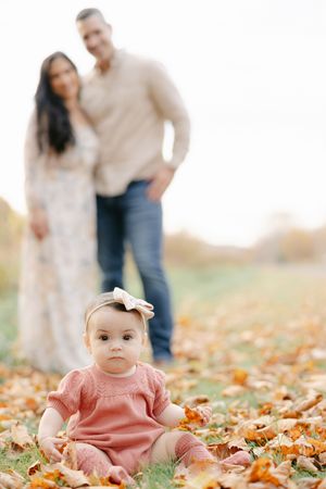 Baby girl in pink dress sits on fall leaves while blurred parents stand together in an autumn outdoor family portrait.