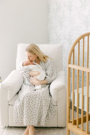 A mother in a floral dress sits in a white nursery chair, tenderly kissing her newborn baby near a wooden crib.