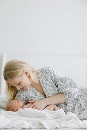 A blonde mother in a floral dress tenderly gazes at her newborn baby wrapped in white blanket on a bed.