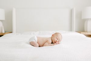 A peaceful newborn baby sleeping on a white bed wearing only a diaper in a bright, minimalist bedroom.