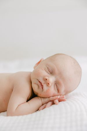 A peaceful newborn baby sleeping on a white blanket with hands tucked under cheek in a serene portrait.