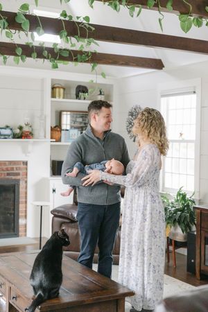 A couple with a newborn baby stands in a cozy living room with exposed beams, a black cat nearby.