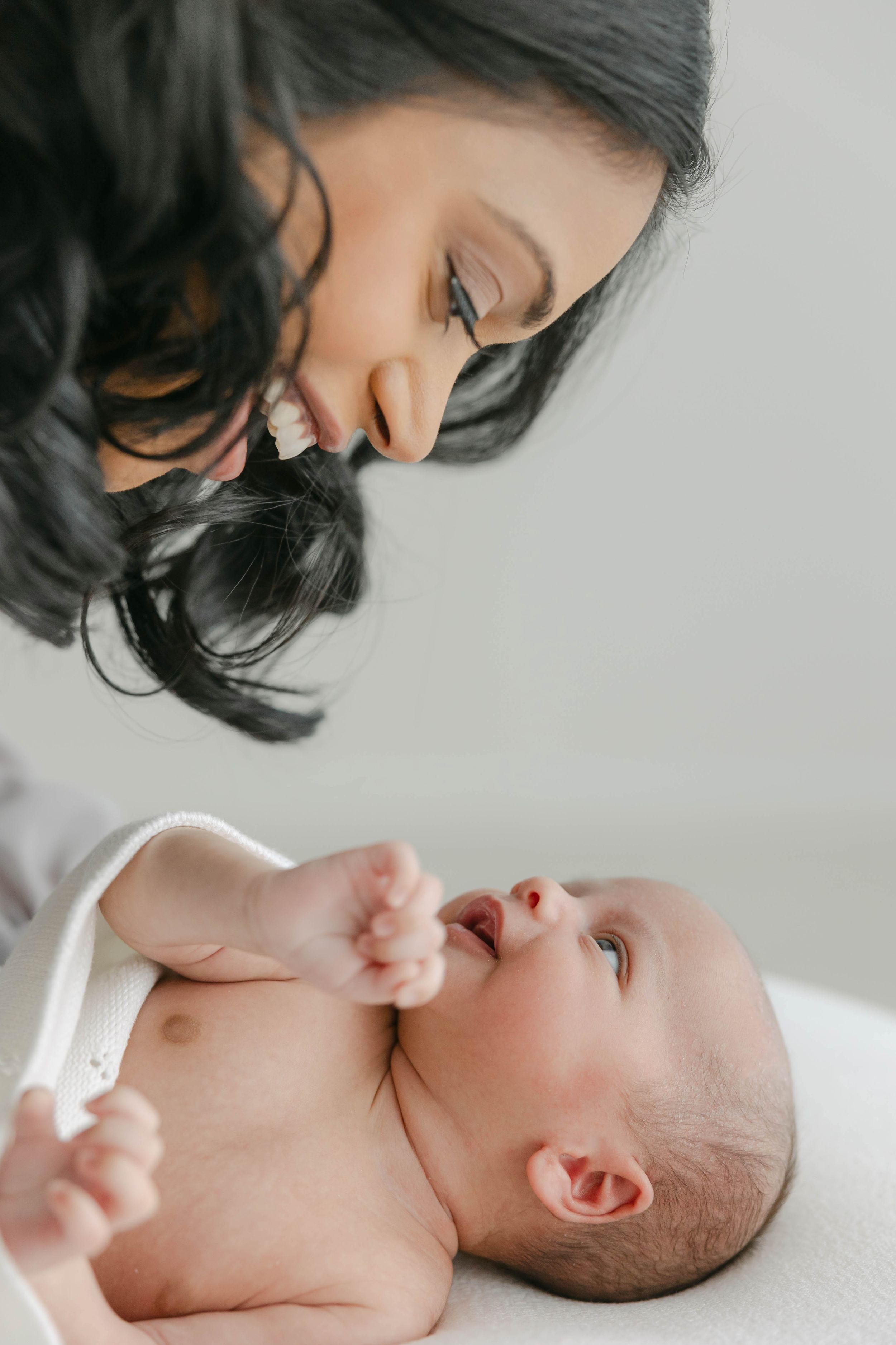 A loving mother with dark curly hair gazes tenderly at her newborn baby lying on a white surface.