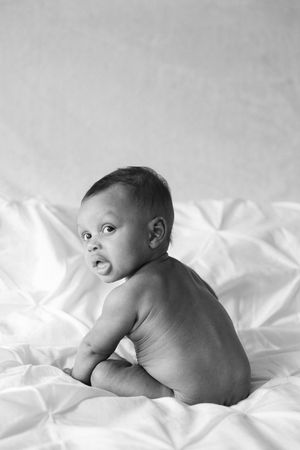 Black and white photo of a cute baby sitting on white bed sheets, looking back over shoulder with curious expression.