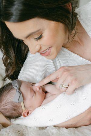 Smiling brunette mother in white gently touches her swaddled newborn baby's hand while bonding closely.